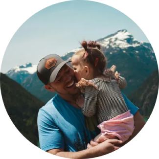 Man with a cap joyfully holding a child, with mountains and forests in the background.