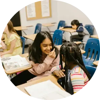 Two young girls smiling and chatting at a classroom desk, practicing communication skills.