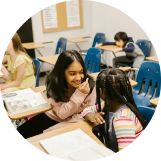 Two young girls smiling and chatting at a classroom desk, practicing communication skills.