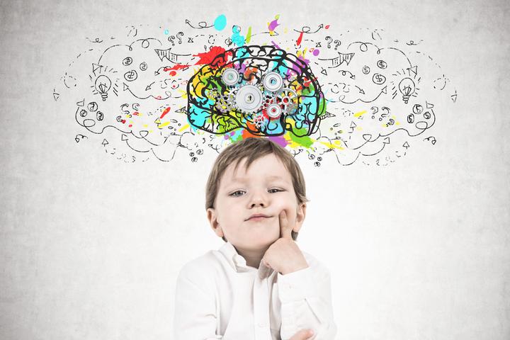 Cute little boy in a white shirt and dark blue jeans is thinking holding his finger near his cheek. Decision making concept. A concrete wall background with a cog brain sketch on it.