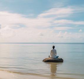 Woman meditating on a rock by the beach facing the water